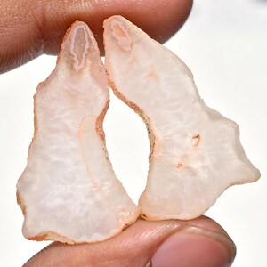 May include: Two raw, natural agate slices. The agate has a translucent white interior with a light peach-colored outer edge. The edges have a rough, uneven texture. The slices are held between fingers against a white background.