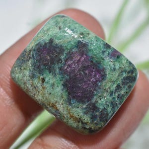 Pode incluir: Uma pedra preciosa polida com formato retangular. A pedra apresenta uma mistura de tons verdes, roxos e vermelho escuro. A superfície tem um acabamento ligeiramente brilhante, destacando os padrões e cores naturais.