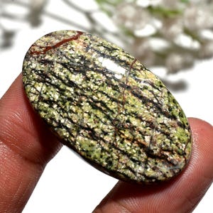 Pode incluir: Uma pedra preciosa oval com um padrão único. A pedra apresenta uma mistura de cores verdes, pretas e castanhas, com uma aparência texturizada. A pedra preciosa é segurada na mão, mostrando sua superfície polida e sua beleza natural. A pedra mede aproximadamente 5 cm de comprimento.