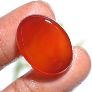 Puede incluir: Una gema pulida, de forma ovalada, en tonos naranja y rojo, se sostiene entre dos dedos. La gema tiene una superficie lisa y brillante, que refleja la luz. El fondo está borroso, destacando el color y la forma de la gema.