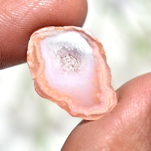 Op de afbeelding: Een gepolijste, ovaalvormige agaatsteen met een centrale kristallijne holte. De steen heeft een kleurengradiënt van wit, roze en perzik, met een doorschijnend uiterlijk. De steen wordt tussen twee vingers gehouden.