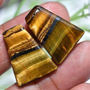 May include: Two polished tiger's eye gemstones. The stones are rectangular with angled edges. The colors are a mix of golden brown, black, and olive green, with a chatoyant effect. The gemstones are held in a hand.