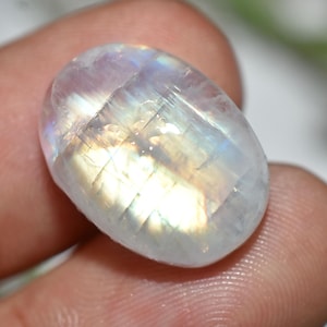 Puede incluir: Una piedra lunar ovalada y facetada. La piedra tiene un color base blanco lechoso con destellos iridiscentes de azul, amarillo y morado. La superficie está pulida y refleja la luz, creando un efecto brillante.