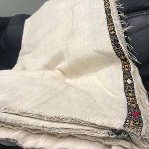 Op de afbeelding: Crèmekleurig geweven textiel met een decoratieve rand. De rand heeft een zwart-goud patroon met accenten van paars, roze en wit. Het textiel is gevouwen en lijkt gemaakt van een zachte, natuurlijke vezel.