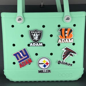 Op de afbeelding: Een lichtgroene draagtas met diverse teamlogo-bedels. De tas heeft een handvat en is versierd met bedels met de logo's van de Raiders, Bengals, Falcons, Steelers en NY, elk met de naam "ADAM" of "MILLER".