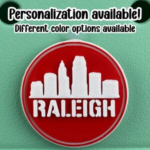 May include: A round, red and white emblem featuring the Raleigh, North Carolina skyline. The word "RALEIGH" is displayed in white block letters below the skyline. The text "Personalization available! Different color options available" is at the top.