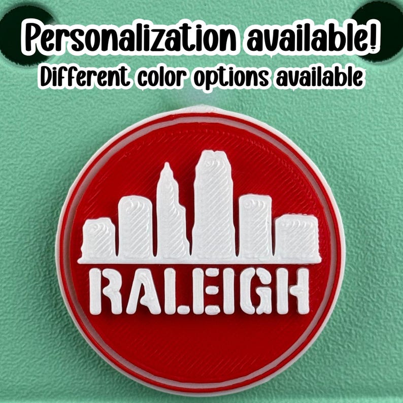 May include: A round, red and white emblem featuring the Raleigh, North Carolina skyline. The word "RALEIGH" is displayed in white block letters below the skyline. The text "Personalization available! Different color options available" is at the top.