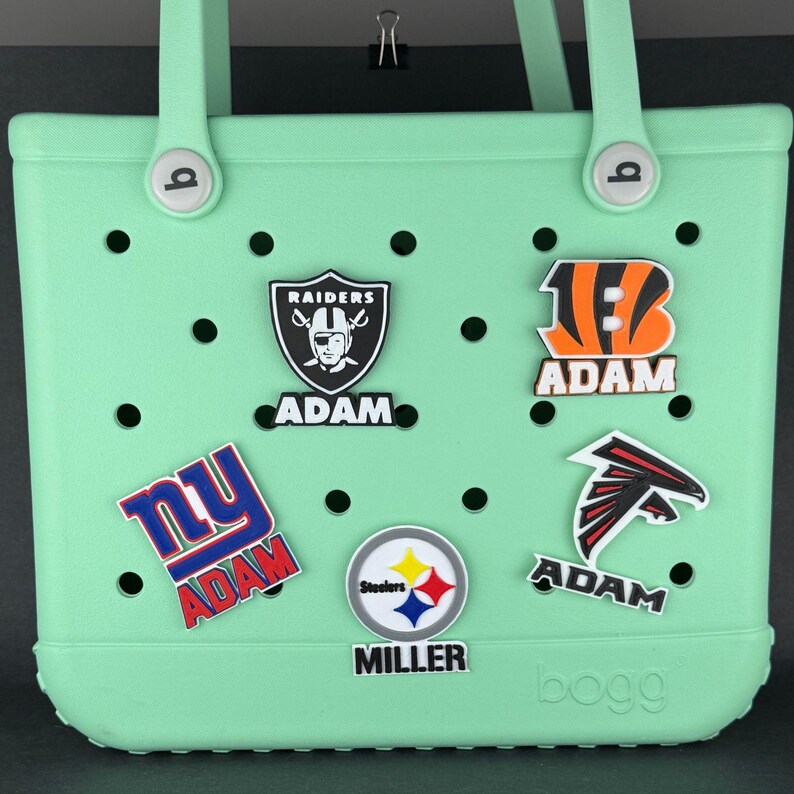 Peut inclure: Un sac fourre-tout vert clair avec plusieurs breloques de logos d'&eacute;quipes. Le sac a une poign&eacute;e et est d&eacute;cor&eacute; de breloques avec les logos Raiders, Bengals, Falcons, Steelers et NY, chacun avec le nom "ADAM" ou "MILLER".