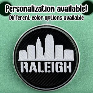 May include: A round, black and white emblem featuring the skyline of Raleigh, North Carolina. The word "RALEIGH" is printed in white block letters below the skyline. The text "Personalization available! Different color options available" is at the top.