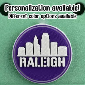 May include: A round purple and white emblem featuring the city skyline of Raleigh, North Carolina. The word "RALEIGH" is printed in white block letters below the skyline. The text "Personalization available! Different color options available" is printed above the emblem.