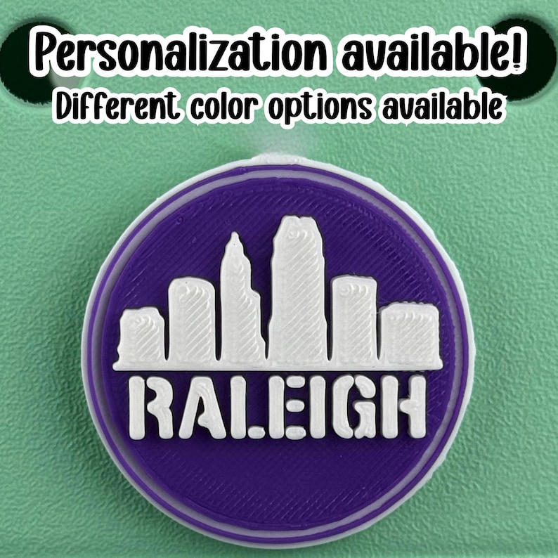 May include: A round purple and white emblem featuring the city skyline of Raleigh, North Carolina. The word "RALEIGH" is printed in white block letters below the skyline. The text "Personalization available! Different color options available" is printed above the emblem.