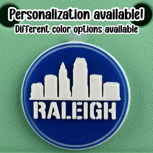 May include: A round, blue and white emblem featuring the Raleigh, North Carolina skyline. The word "RALEIGH" is printed in white block letters below the skyline. The text "Personalization available! Different color options available" is printed above the emblem.