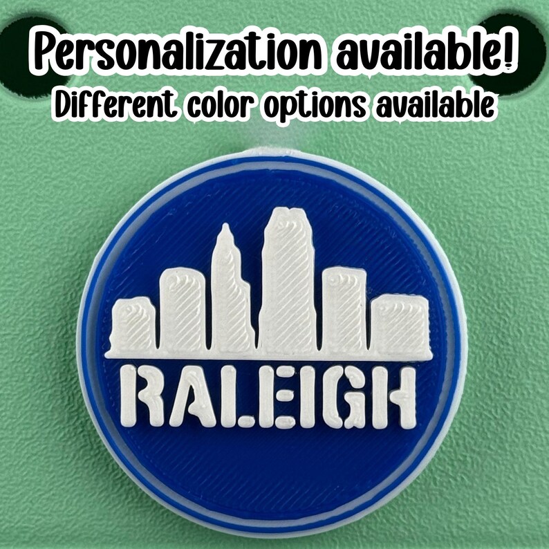 May include: A round, blue and white emblem featuring the Raleigh, North Carolina skyline. The word "RALEIGH" is printed in white block letters below the skyline. The text "Personalization available! Different color options available" is printed above the emblem.
