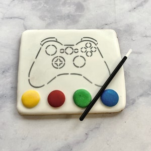 May include: A white cookie decorated with a black outline of a video game controller. The cookie has four small circles of icing in yellow, red, green, and blue. A black paintbrush is included.