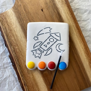 May include: A white cookie decorated with a black and white outline of a rocket ship, stars, and a crescent moon. The cookie has four colorful candies on the bottom, resembling a paint palette. A black paintbrush is lying next to the cookie.
