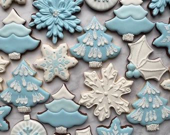 Puede incluir: Una colección de galletas decoradas con glaseado real en tonos azul claro y blanco. Las formas incluyen copos de nieve, árboles de Navidad, adornos redondos y hojas de acebo, con detalles intrincados y acentos de azúcar brillante.