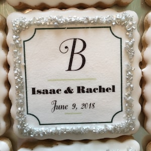 May include: A white cookie with a silver sugar border. The cookie has a printed design with the letter "B", the names "Isaac & Rachel", and the date "June 9, 2018".