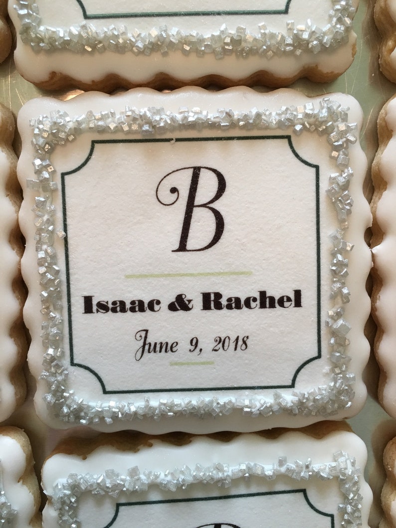 May include: A white cookie with a silver sugar border. The cookie has a printed design with the letter "B", the names "Isaac & Rachel", and the date "June 9, 2018".