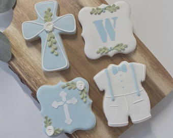Op de afbeelding: Vier versierde koekjes bestaande uit een lichtblauw kruis met wit bloemmotief, een wit vierkant koekje met een lichtblauwe letter W, een lichtblauw vierkant koekje met een wit kruis en een koekje in de vorm van een outfit met bretels en strik.