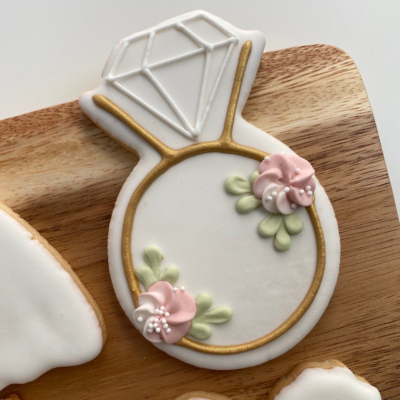 May include: A white sugar cookie decorated with gold icing in the shape of a ring with a diamond on top. The ring is decorated with pink and green icing flowers.