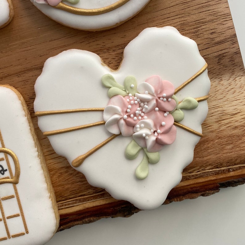 May include: A white heart-shaped cookie decorated with gold lines and a cluster of pink and white flowers with green leaves. The flowers are accented with small white sprinkles.