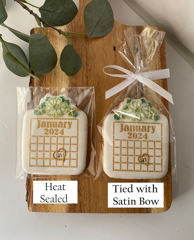 May include: Two white sugar cookies decorated with green floral designs and gold lettering. The cookies are shaped like calendars and say "January 2024". One cookie is heat sealed in a clear plastic bag, while the other is tied with a white satin ribbon in a clear plastic bag.