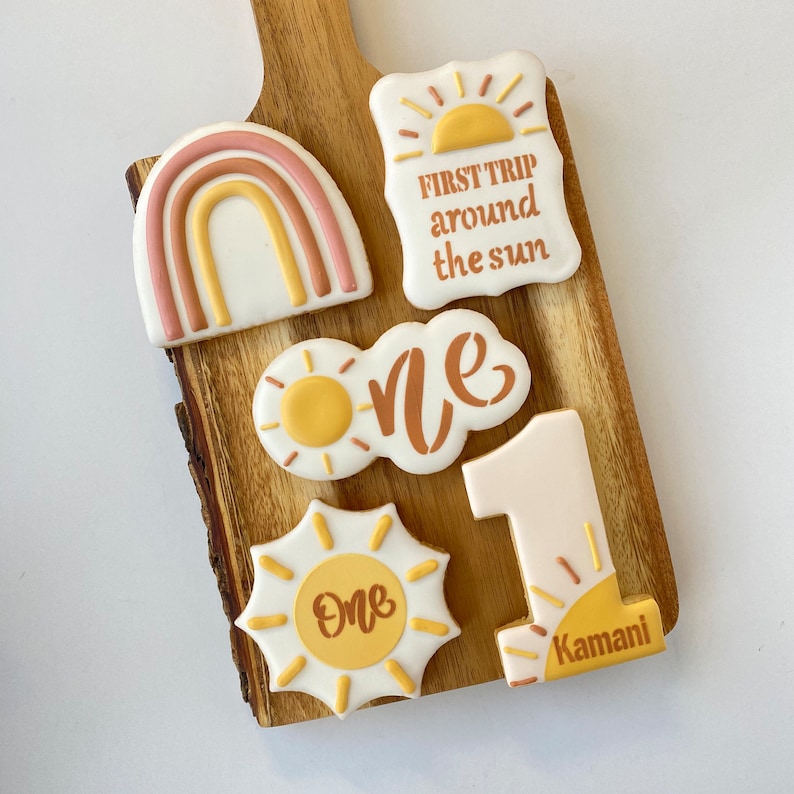May include: A set of six decorated sugar cookies with a sun theme. The cookies are decorated with yellow, orange, and brown icing. The cookies feature the words "First Trip Around the Sun", "One", and "Kamani".