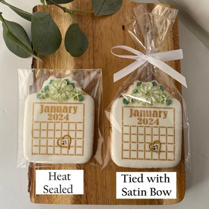 May include: Two white sugar cookies decorated with green floral designs and gold lettering. The cookies are shaped like calendars and say "January 2024". One cookie is heat sealed in a clear plastic bag, while the other is tied with a white satin ribbon in a clear plastic bag.