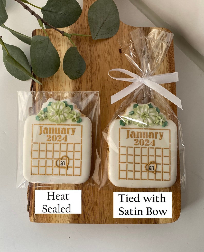 May include: Two white sugar cookies decorated with green floral designs and gold lettering. The cookies are shaped like calendars and say "January 2024". One cookie is heat sealed in a clear plastic bag, while the other is tied with a white satin ribbon in a clear plastic bag.