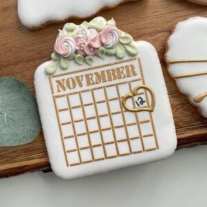 May include: A white sugar cookie decorated with gold icing in the shape of a calendar with the word "NOVEMBER" and the number "12" inside a heart. The cookie is topped with a cluster of pink and green icing flowers.