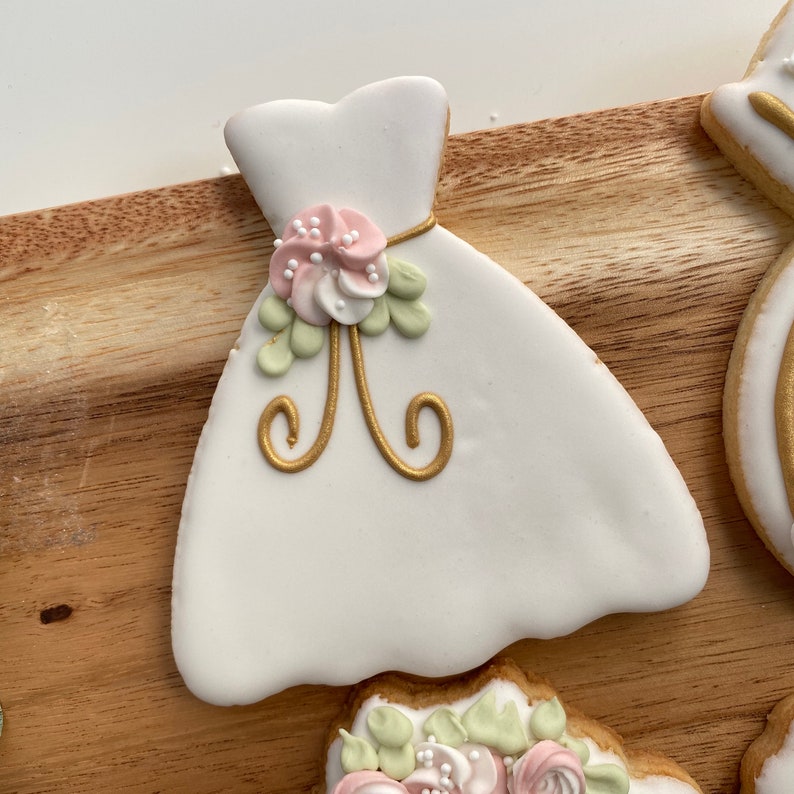 May include: White sugar cookie decorated to look like a wedding dress. The dress has a gold ribbon with a pink and green floral design.