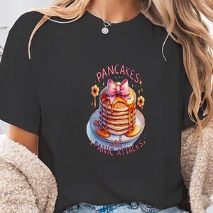 Könnte beinhalten: Schwarzes T-Shirt mit einem Pfannkuchen-Design, Sirup und einer rosa Schleife. Der Text lautet "Pancakes with Panic Attacks". Das Design umfasst gelbe Blumenakzente und einen weißen Teller. Ein Freizeitkleidungsstück.