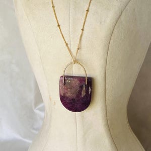 Könnte beinhalten: Eine goldfarbene Halskette mit einem Anhänger aus einem halbkreisförmigen, violetten Stein. Der Stein hat ein marmoriertes Aussehen mit Rosa- und Lilatönen. Die Halskette wird auf einer Schaufensterpuppe präsentiert.