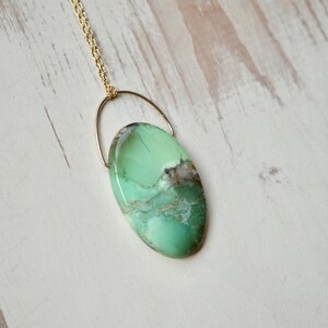 Puede incluir: Un collar de cadena dorada con un colgante ovalado verde. El colgante est&aacute; hecho de una piedra verde con vetas blancas y marrones.