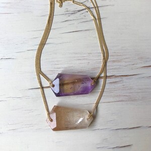 May include: A gold chain necklace with two crystal pendants. The top pendant is a purple amethyst crystal and the bottom pendant is a clear quartz crystal.