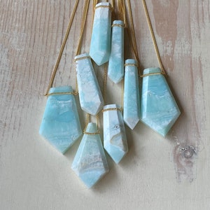 Pu&ograve; includere: Collezione di collane con pendenti in pietra preziosa azzurra e bianca. Ogni pendente ha una forma unica ed &egrave; sospeso a una delicata catena d'oro. Le collane sono esposte su uno sfondo di legno chiaro.
