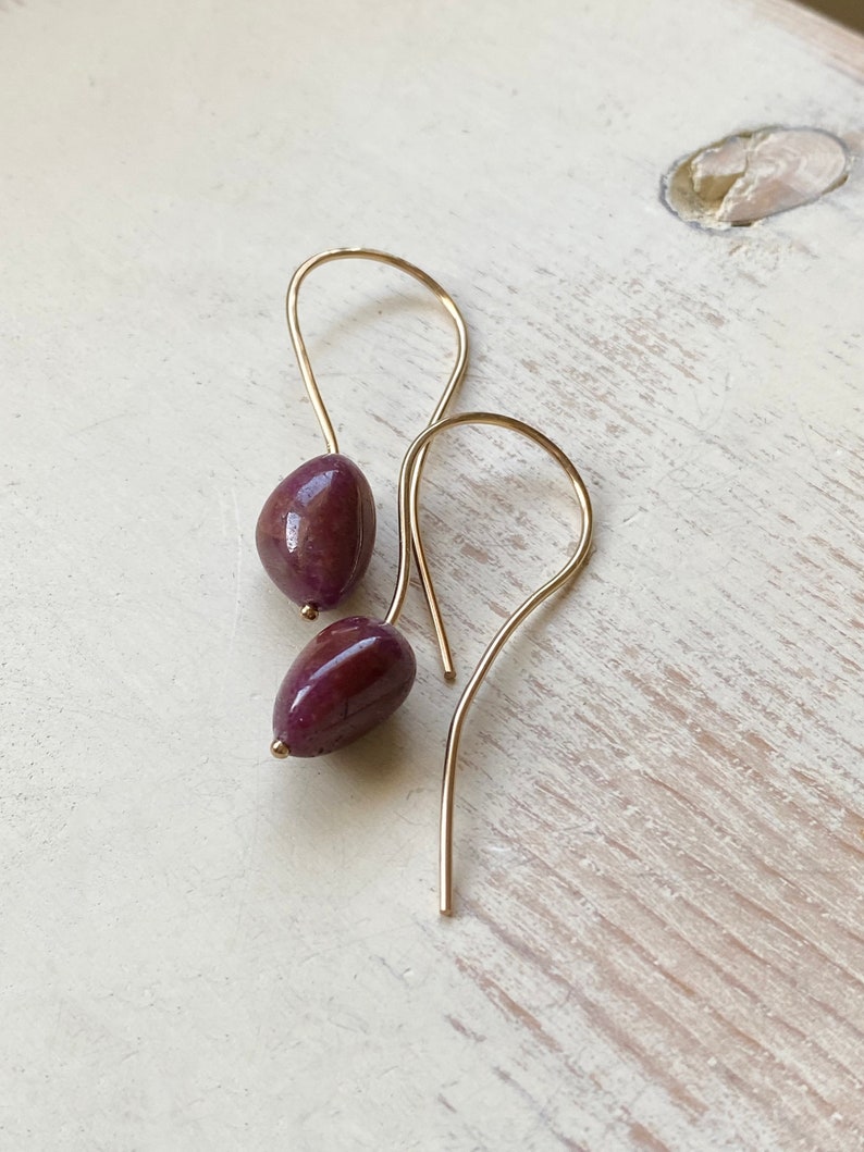 May include: A pair of gold earrings with a teardrop-shaped purple gemstone hanging from each.