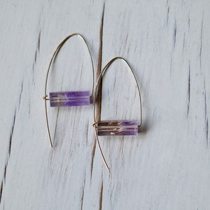 May include: A pair of gold-colored wire earrings, each featuring a rectangular, translucent purple gemstone. The earrings are displayed against a white, distressed wooden surface, showcasing their minimalist design and natural stone accents.