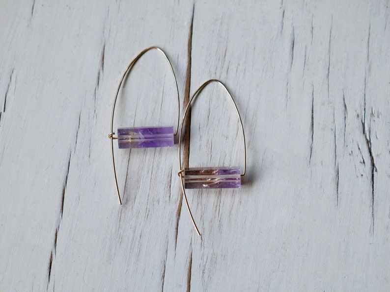 May include: A pair of gold-colored wire earrings, each featuring a rectangular, translucent purple gemstone. The earrings are displayed against a white, distressed wooden surface, showcasing their minimalist design and natural stone accents.