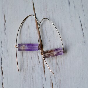 May include: A pair of earrings featuring rectangular amethyst beads suspended from gold-colored wire. The amethyst stones display a mix of purple and clear hues, creating a striking contrast with the gold wire. The earrings are displayed against a white wooden surface.