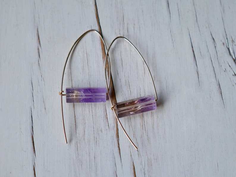 May include: A pair of earrings featuring rectangular amethyst beads suspended from gold-colored wire. The amethyst stones display a mix of purple and clear hues, creating a striking contrast with the gold wire. The earrings are displayed against a white wooden surface.