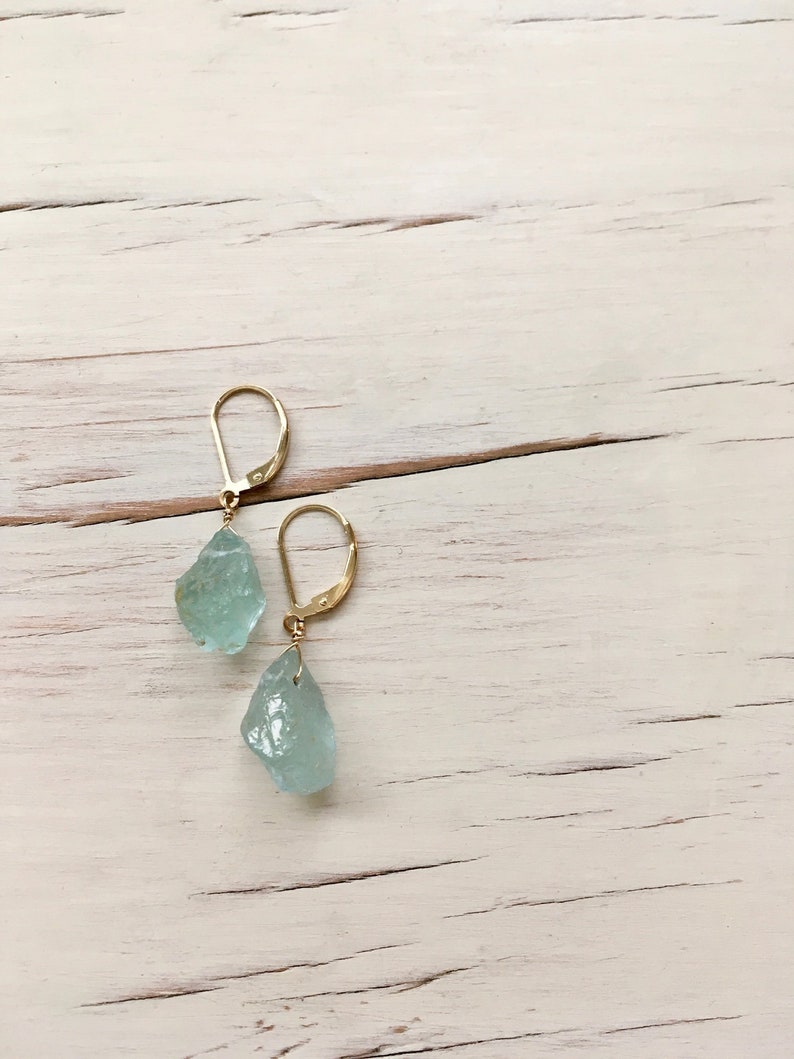 Peut inclure: Une paire de boucles d'oreilles en or avec des pierres d'aigue-marine bleu clair. Les pierres sont brutes et ont une forme naturelle irr&eacute;guli&egrave;re.