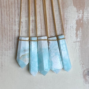 Pu&ograve; includere: Cinque collane con pendenti in cristallo azzurro chiaro. Ogni pendente ha una forma esagonale allungata, sospeso a una catena dorata. I cristalli hanno un aspetto marmorizzato con sfumature bianche e azzurre. Le collane sono esposte su uno sfondo di legno chiaro.