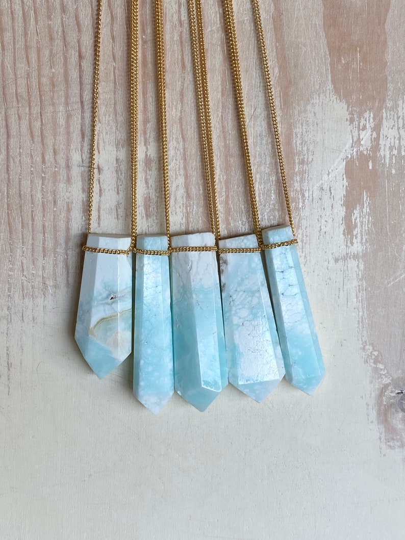 Pu&ograve; includere: Cinque collane con pendenti in cristallo azzurro chiaro. Ogni pendente ha una forma esagonale allungata, sospeso a una catena dorata. I cristalli hanno un aspetto marmorizzato con sfumature bianche e azzurre. Le collane sono esposte su uno sfondo di legno chiaro.