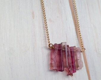 Könnte beinhalten: Eine goldene Kettenkette mit einem Anhänger aus mehreren rosa und klaren Turmalin-Steinen. Die Steine sind rechteckig geschliffen und in einer Traube angeordnet.