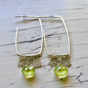 May include: A pair of silver hoop earrings with a dangling chain and a green gemstone. The earrings are on a white wooden surface.