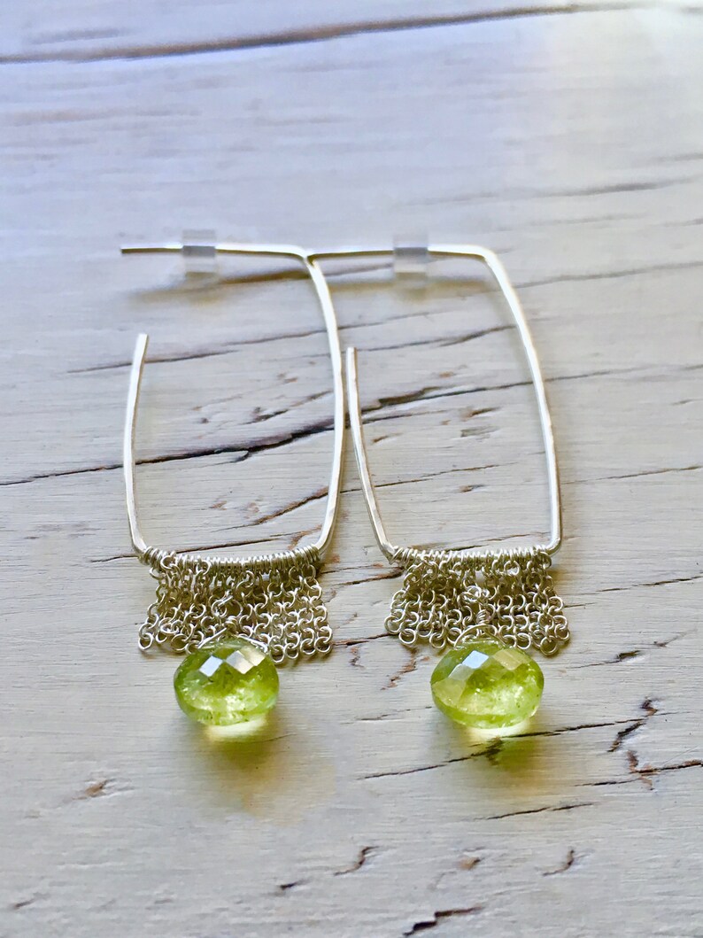 May include: A pair of silver hoop earrings with a dangling chain and a green gemstone. The earrings are on a white wooden surface.