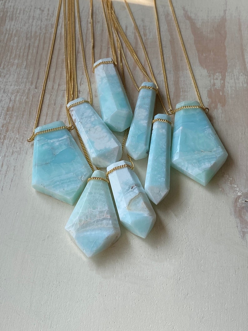 Pu&ograve; includere: Una collezione di collane con pendenti in pietre preziose e catene dorate. I pendenti, di varie forme e dimensioni, presentano una combinazione di colori azzurro chiaro e bianco. Le collane sono esposte su una superficie di legno chiaro.