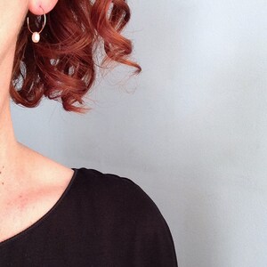 May include: A woman with auburn hair wearing a pair of small gold hoop earrings with a single pearl dangling from each hoop.