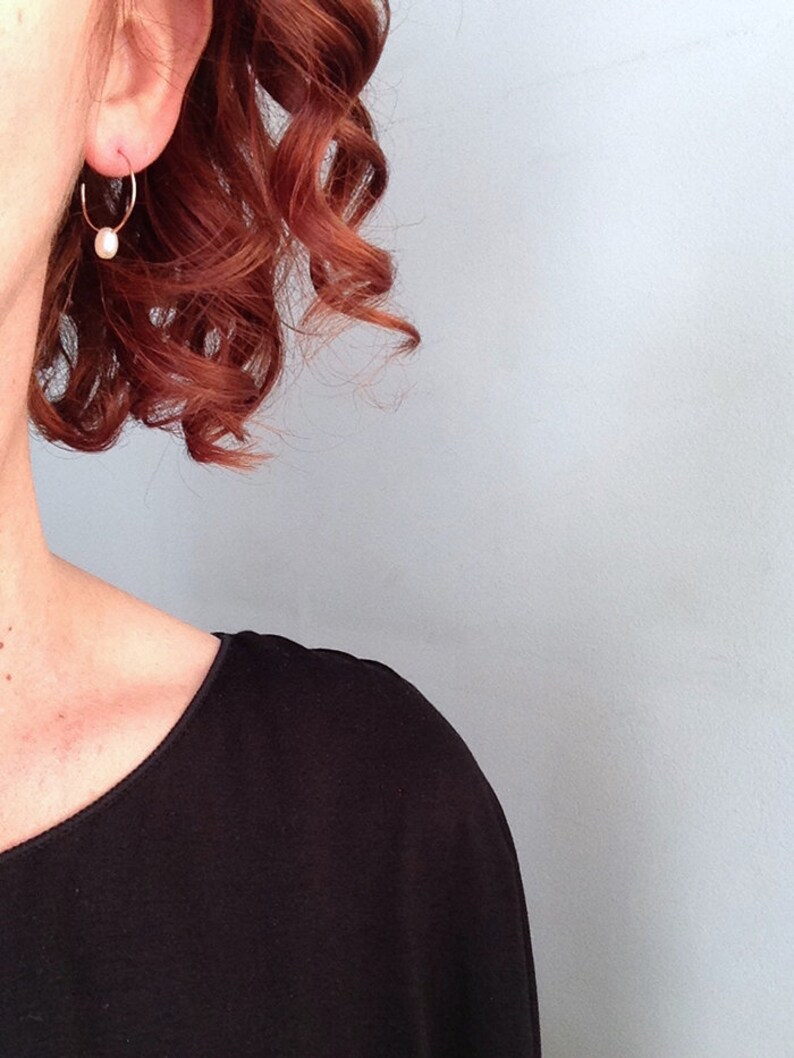 May include: A woman with auburn hair wearing a pair of small gold hoop earrings with a single pearl dangling from each hoop.